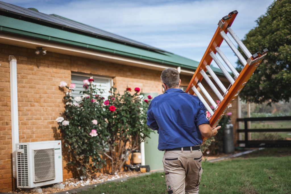A person in a blue shirt and beige pants carries an orange ladder towards a brick house with solar panels on the roof and flowers by the window. An air conditioning unit is mounted on the exterior wall.