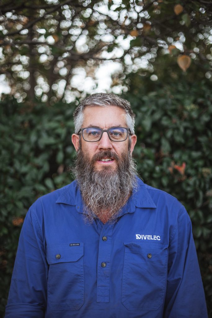 A man with a long gray beard and glasses is wearing a blue shirt with "DIVELEC" embroidered on it. He is standing outdoors in front of a leafy background.