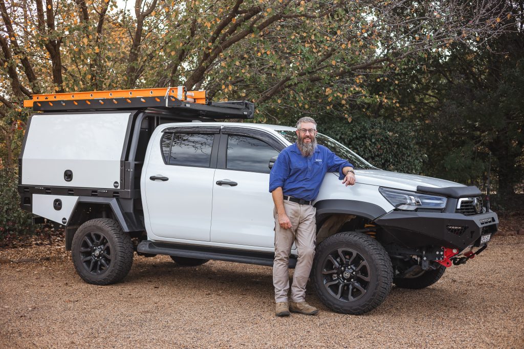 A man with a beard wearing a blue shirt and khaki pants stands next to a white off-road vehicle with a roof rack and storage compartment, parked on a gravel surface with trees in the background.
