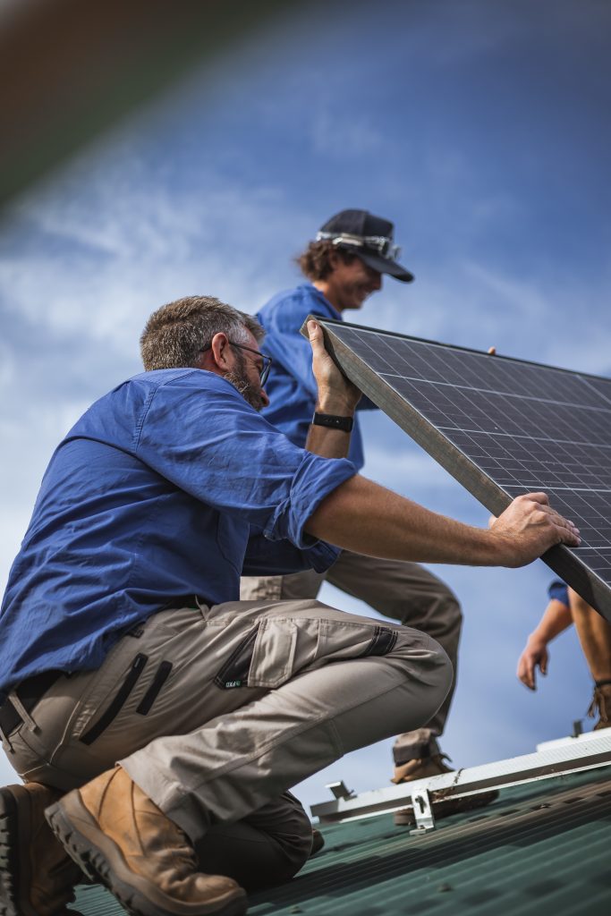 Two workers wearing blue shirts and safety gear are installing a solar panel on a rooftop under a blue sky.