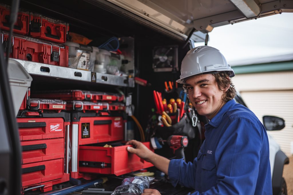 A person wearing a hard hat and blue shirt smiles while organizing red toolboxes and various tools in a utility vehicle.