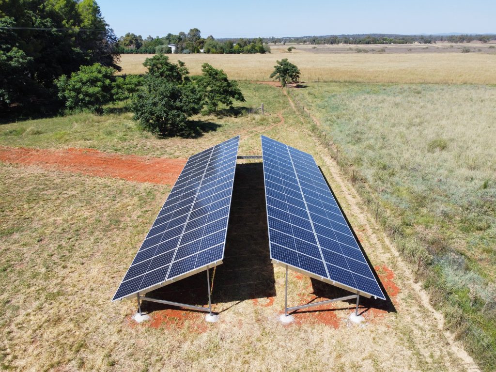 Two arrays of solar panels installed in a rural field, surrounded by grass and trees, on a sunny day.