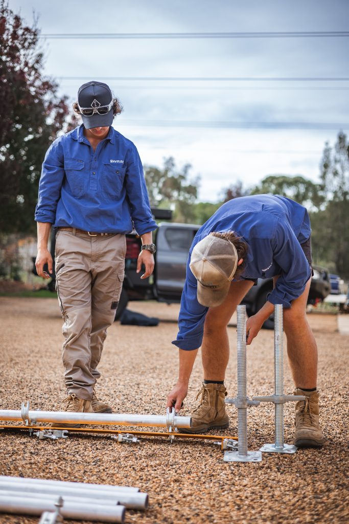 Two individuals in blue shirts and work pants are assembling metal frames outdoors on a gravel surface. One person is securing a joint while the other stands nearby, observing the process.