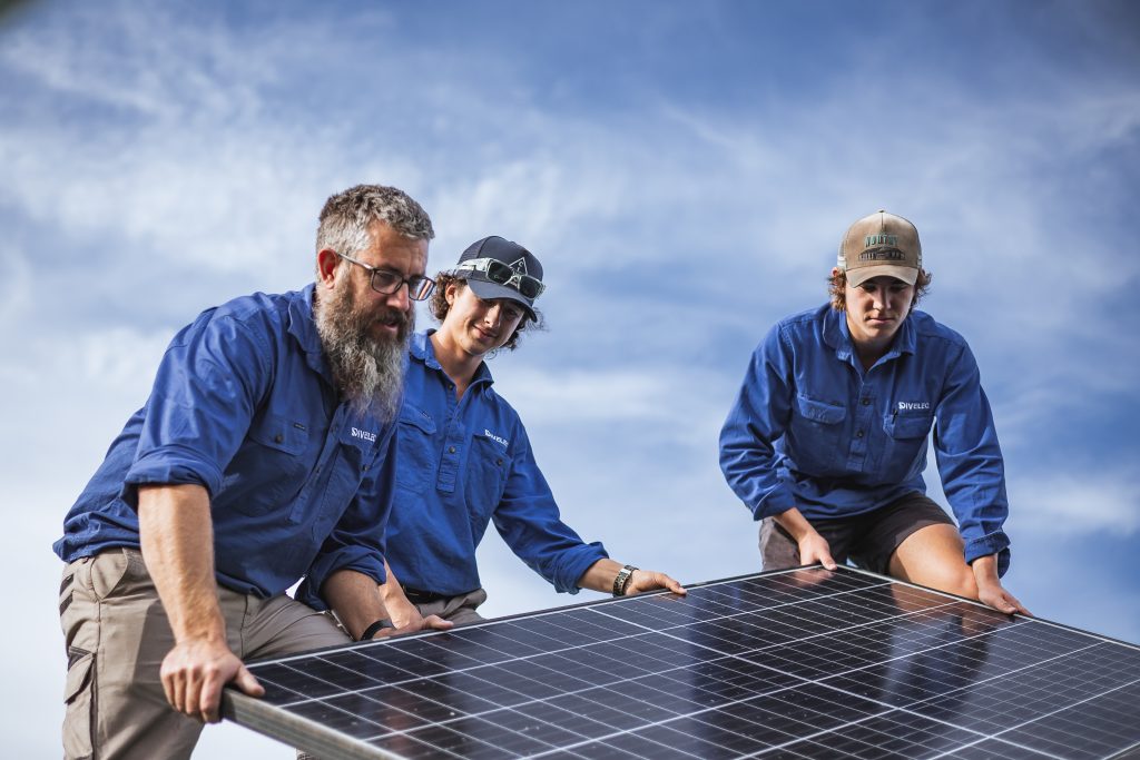 Three individuals in blue shirts are installing a solar panel under a partly cloudy sky.