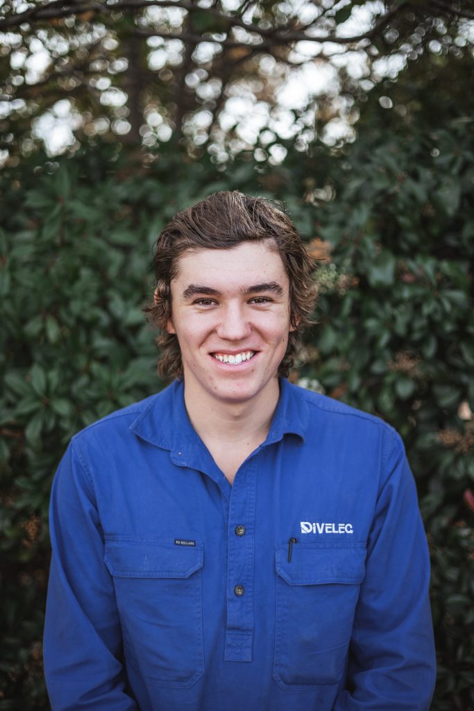 A person wearing a blue shirt smiles while standing outdoors with greenery in the background.