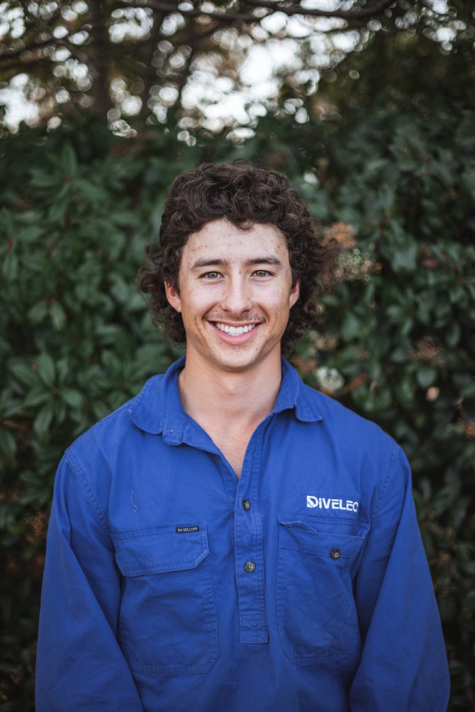 A man with curly hair, wearing a blue shirt with the logo "DIVELCO" stands in front of a leafy background, smiling at the camera.