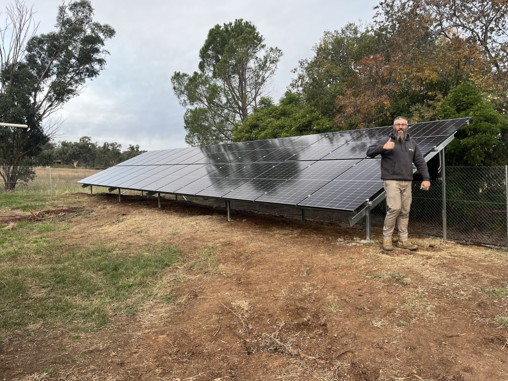 A man stands next to a ground-mounted solar panel array in a grassy area, giving a thumbs-up. Trees are visible in the background.