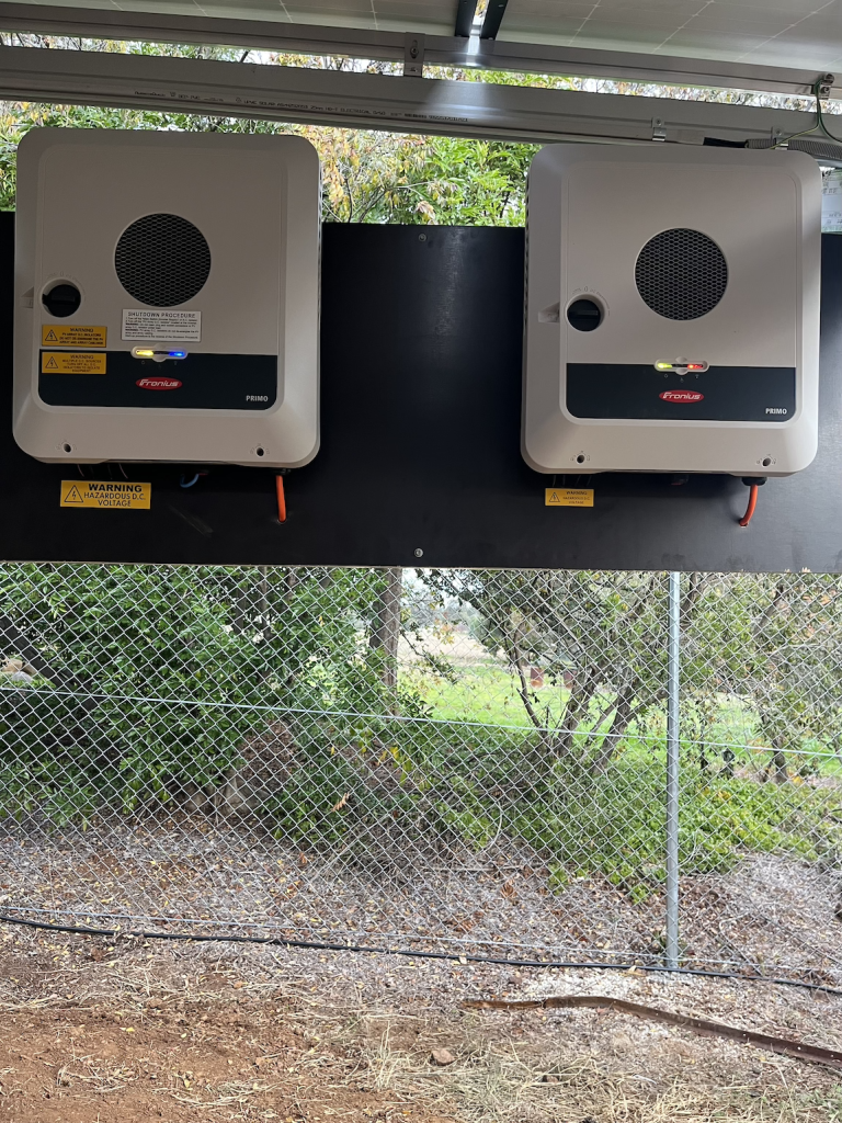 Two inverter units mounted on a metal board, with a wire fence and green foliage in the background. Safety warning labels are visible on the inverters.