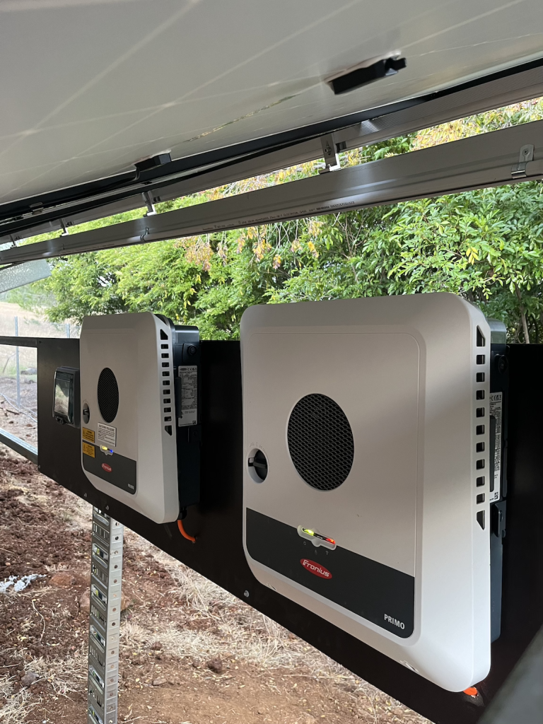 Two electrical devices mounted side-by-side on a black panel, set outdoors near green foliage. The devices are rectangular and feature ventilation grilles.