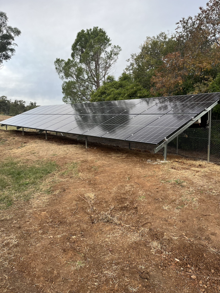 A ground-mounted solar panel system installed in a grassy area with trees in the background on a cloudy day.