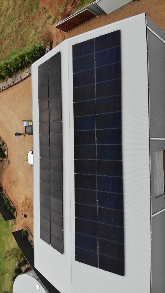 An aerial view of a building with a gray roof that has two large arrays of solar panels. The surrounding area features a gravel driveway, grassy patches, and a person near a vehicle.
