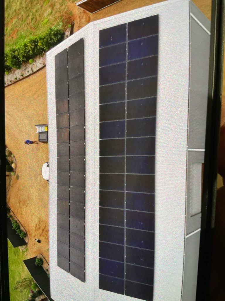 Aerial view of a building with two rows of solar panels on the roof, surrounded by a grassy area and a person standing near a small structure.