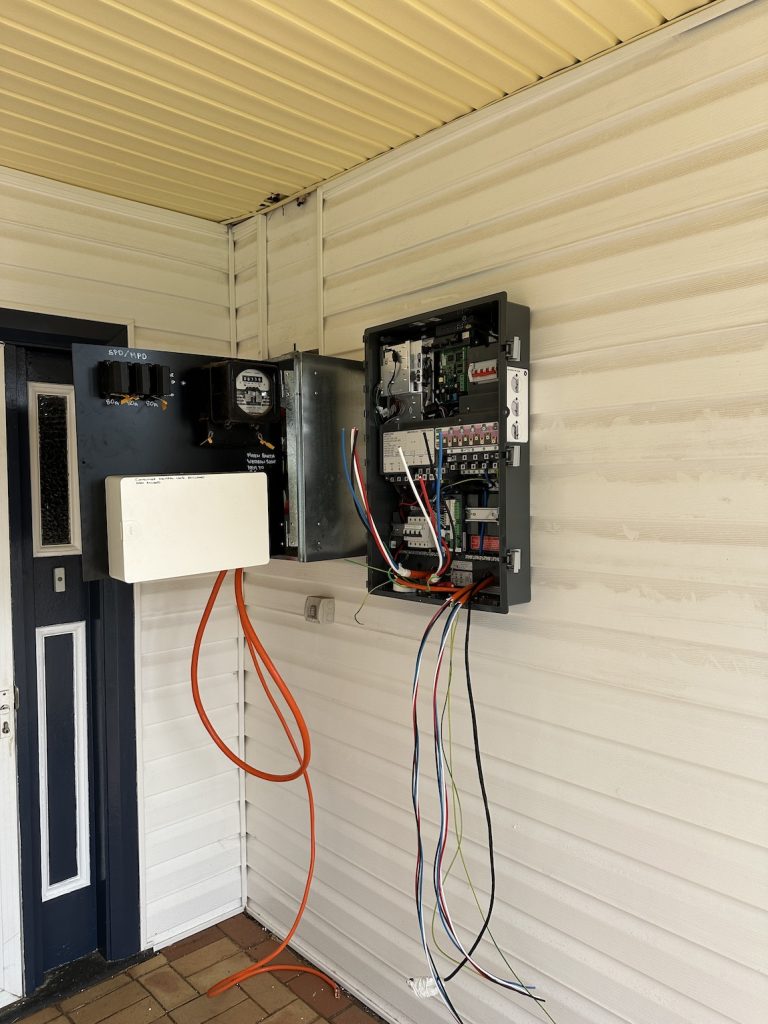 A maintenance room with an open electrical panel and several exposed wires running through it. The panel is installed on a beige paneled wall.