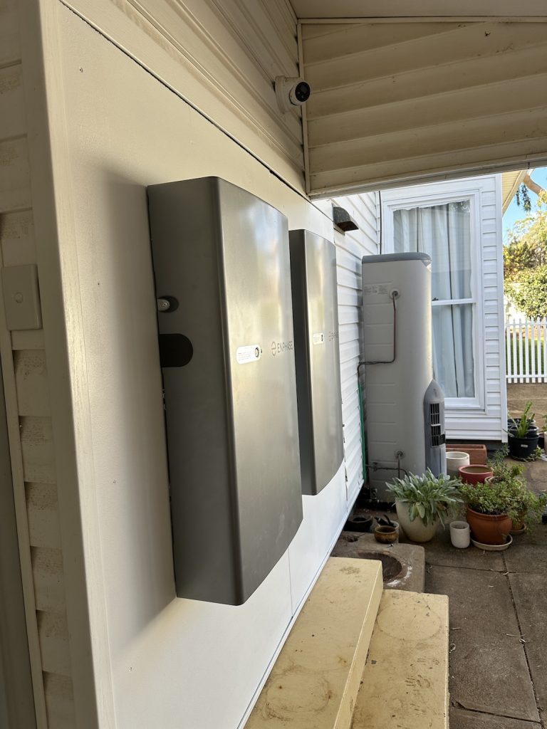 Two wall-mounted metal electrical boxes, a rainwater tank, and several potted plants are on the porch of a white house with a window and a security camera overhead.