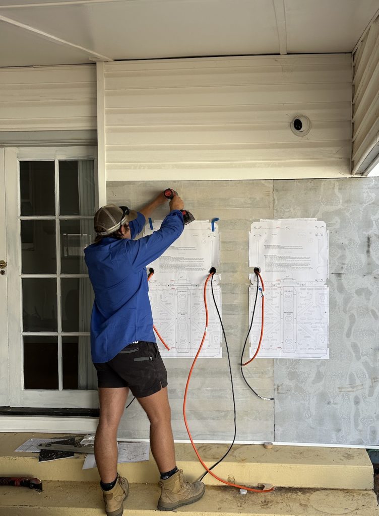 A person in work attire uses a power drill on a wall with two large sheets of paper taped to it. Electric cords are connected to the wall.